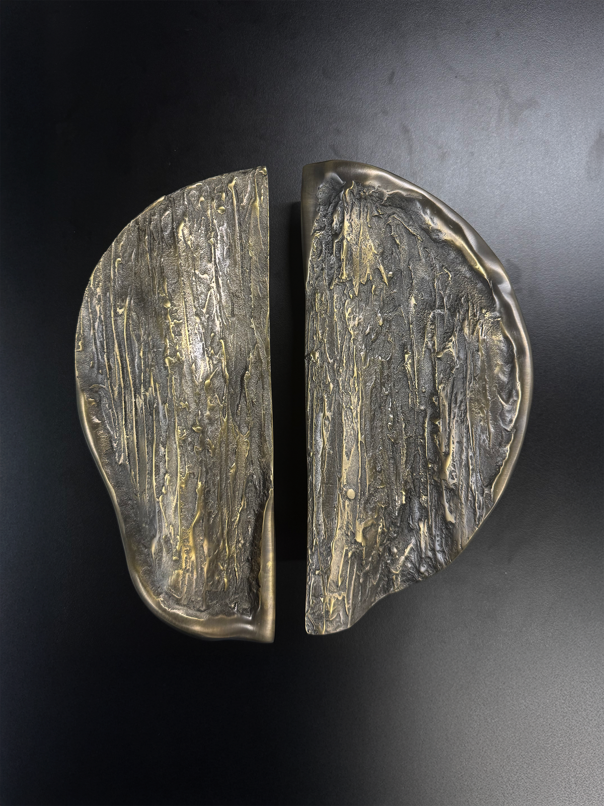 A pair of large, organic-shaped metallic door handles with a textured, hammered finish in a brushed gold and dark bronze tone, shown on a dark background.